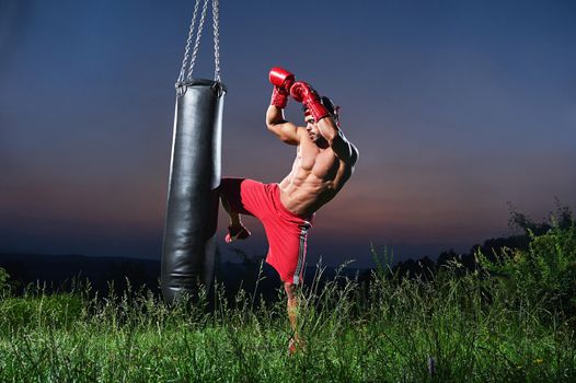 Handsome shirtless muscular young kick boxer working out with a punching bag outdoors copyspace beautiful sunset on the background nature lifestyle sports active athlete athletic masculinity training.