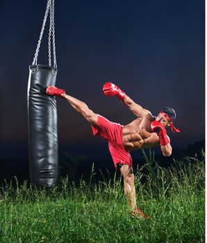 Handsome young strong muscular fighter exercising with a punching bag practicing his high kick martial arts combat fighting kickboxing Muay Thai strength power masculinity energy effort athlete.
