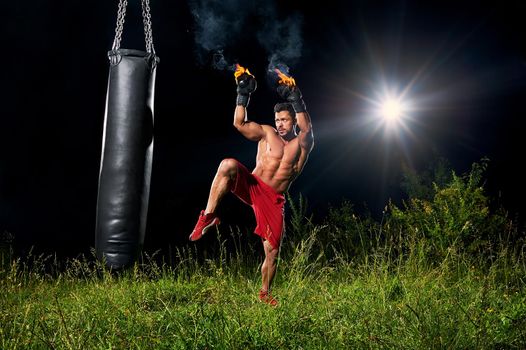 Full length shot of a handsome young muscular male fighter wearing boxing gloves burning with fire training on a punching bag outdoors at night copyspace athletics sports fitness martial arts combat.