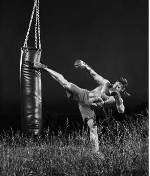 Full length monochrome shot of a muscular young kick boxer kicking a heavy bag training outdoors nature workout exercising combat martial fitness sport sportive sportsman athletics physique.