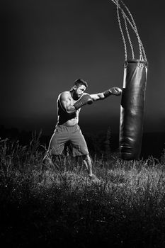 Vertical monochrome shot of a young male boxer with ripped strong muscular body hitting a punching bag working out outdoors at night professional fighter fighting boxing technique skills fitness sport.