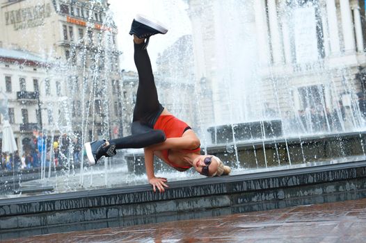 Cool female hip-hop dancer making a handstand while dancing near the fountain at the city center performing outdoors.