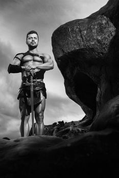 Guarding his world. Monochrome shot of a manly gladiator with strong muscular body looking away thoughtfully standing on a rock