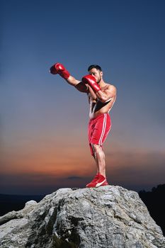 Focused young male boxer training outdoors wearing boxing gloves working out on sunset punching fighting muscles power strength confidence defense martial sports sportive body sexy handsome guy.