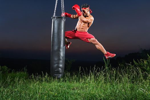 Professional kick boxer jumping and kicking a punching bag with his knee training outdoors on sunset copyspace workout exercising practicing performance preparing competitive motivated