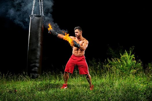 Young handsome muscular man wearing burning boxing gloves punching a heavy sandbag working out at night outdoors copyspace sport fitness martial combat fighting fighter strength effort energy.
