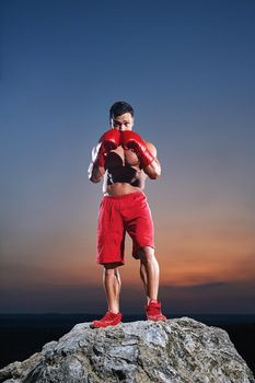 Portrait of a fit young muscular man wearing boxing gloves training outdoors on sunset punching exercising working out preparing competitive motivated determined energetic masculine powerful fighter.