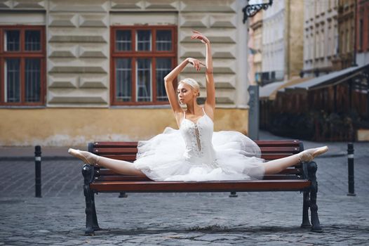 Horizontal shot of a gorgeous blonde haired ballerina doing splits on the bench in the city center.