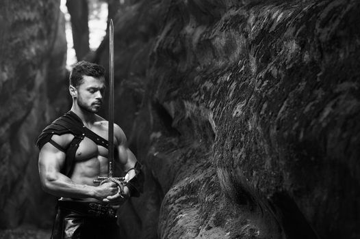 Power in peace. Monochrome shot of a calm thoughtful warrior standing with a sword near the rocks at the woods. Young strong man with a muscular torso posing copyspace