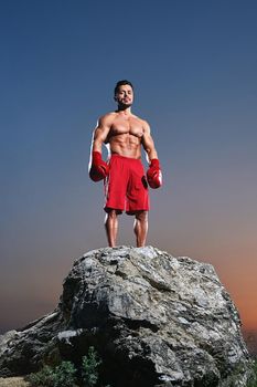 Young male boxer wearing boxing gloves looking to the camera confidently standing on top of a rock on sunset showing off his strong sexy muscular body power masculinity sport sportsperson lifestyle.