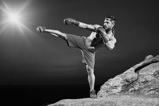 Horizontal full length portrait of a shirtless muscular fighter exercising outdoors kickboxing combat sport sportsman athlete muscular strong effort competitive practicing workout concept monochrome