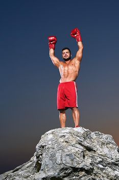 Boxing champion celebration victory standing on a rock with his arms raised in the air shirtless muscular strong competitive first championship fighter sportsman athlete competition winner winning.