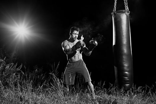 Monochrome portrait of a focused male boxer exercising outdoors hitting a punching bag wearing burning boxing gloves copyspace concentration determination fit toned body torso strengthening concept.
