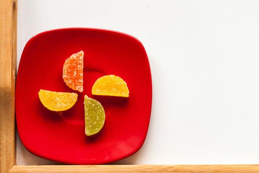 Four Sugar candies. on a red plate. Coloured green, yellow, orange. Shaped as half moon. Placed on a left, down corner.