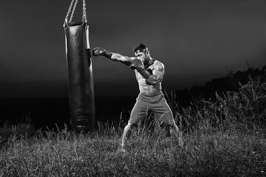 Black and white full length of a shirtless young male boxer practicing boxing technique on a punching bag outdoors copyspace nature concentration determination effort achieving competitive tough.