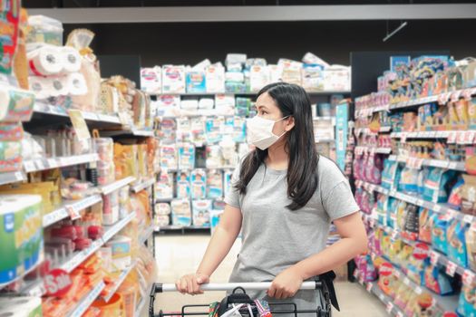Customer Asian Woman Wearing Face Mask With Shopping Cart in Supermarket Department Store Shop While Choosing and Looking Goods on Shelf During Covid-19 Pandemic. Coronavirus Covid Situation