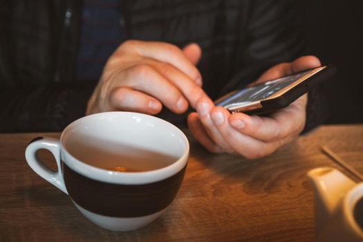 A man is resting in a cafe, drinking tea, reading and writing messages on a smartphone. Work in the mobile office. All-day сommunication. Close-up soft focus image with shallow depth of field