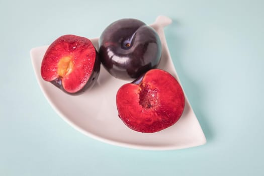 On a white saucer is a large plum, cut in half, and next to it is a whole plum. The pulp of red berries. Close-up.