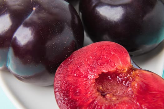 On a white saucer is a large plum, cut in half, and next to it is a whole plum. The pulp of red berries. Close-up.