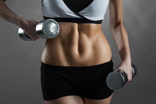 Flat and strong. Cropped closeup of a fitness woman in sports top and shorts holding weights showing off her perfect abs