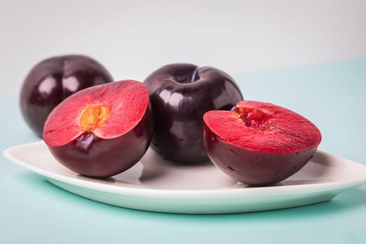 On a white saucer is a large plum, cut in half, and next to it is a whole plum. The pulp of red berries. Close-up.