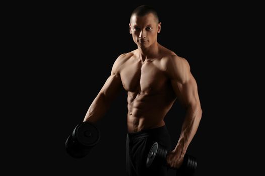 In the name of perfect body. Horizontal studio shot of a young bodybuilder man posing with dumbbells smiling to the camera posing shirtless with his fit and toned torso