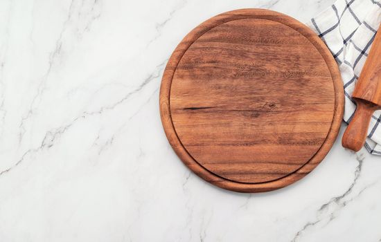 Empty wooden pizza platter with napkin and rolling pin set up on marble stone kitchen table. Pizza board and tablecloth on white marble background.