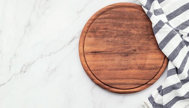 Empty wooden pizza platter with napkin set up on marble stone kitchen table. Pizza board and tablecloth on white marble background.