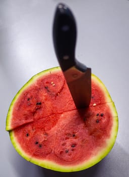 A large knife is stuck in a ripe red watermelon, we will stitch it on a gray table.
