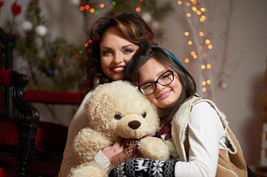 2018. Nothing better than loving home. Young girl hugging a teddy bear enjoying Christmas eve at home with her cheerful mother soft focus and noise