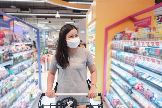 Customer Asian Woman Wearing Face Mask With Shopping Cart in Supermarket Department Store Shop While Choosing and Looking Something on Shelf During Covid-19 Pandemic. Coronavirus Covid Situation