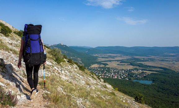 A girl with a large blue backpack climbs a trail in the mountains of the Crimean peninsula.