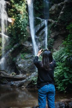 Tourist Woman is Enjoying While Photographing Waterfall on Travel Vacation, Rear View of Asian Woman Relaxing Outdoor Adventure Tourism and Leisure Activity Lifestyle. Holiday Outdoors Trip