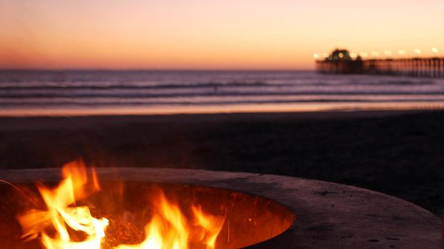 Campfire pit by Oceanside pier, California USA. Camp fire burning on ocean beach, bonfire flame in cement ring place for bbq, sea water waves. Romantic evening twilight sky, dusk after summer sunset.