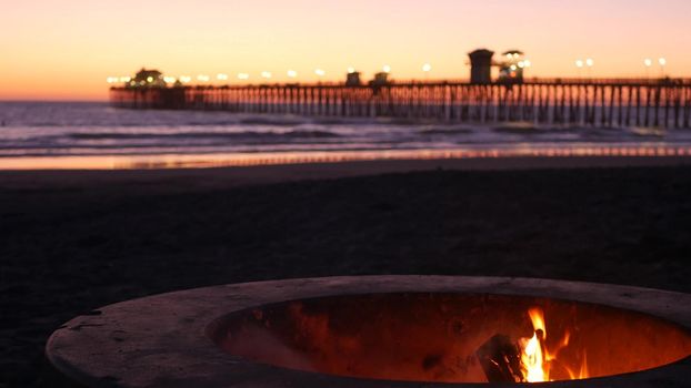 Campfire pit by Oceanside pier, California USA. Camp fire burning on ocean beach, bonfire flame in cement ring place for bbq, sea water waves. Romantic evening twilight sky, dusk after summer sunset.