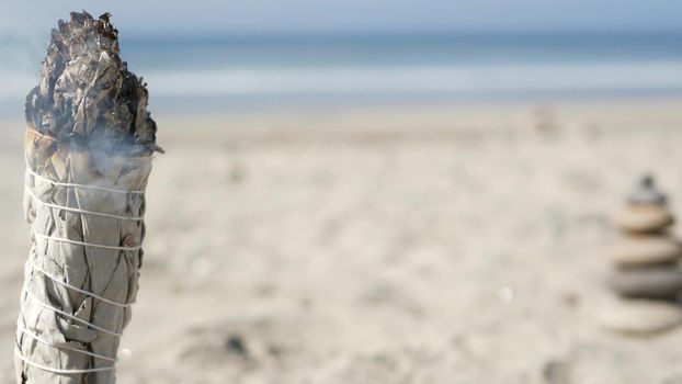 Dried white sage smoke, dry smudge stick burning, soft focus bokeh. Aroma smudging close up on ocean beach, aromatherapy meditation by sea water waves on sandy coast. Yoga, peace, harmony and ayurveda