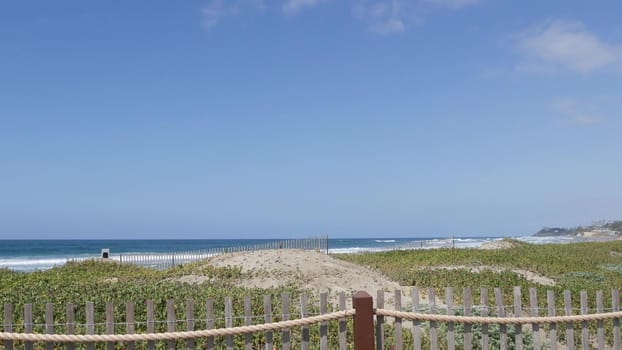 Blue turquoise water surf, big tide waves on sunny summer beach, Encinitas shoreline, California USA. Pacific ocean coast, greenery and wooden picket fence on sea shore. Coastline near Los Angeles.