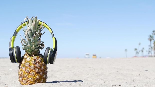 Funny pineapple in headphones, sandy ocean beach, yellow lifeguard car, California USA, Pacific coast. Tropical summer exotic fruit enjoying vacations and music in paradise. Ananas sunbathing on shore