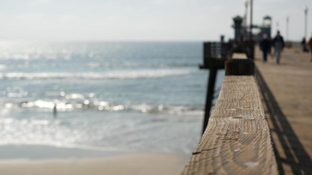 Railings of old wooden pier, people walking on waterfront boardwalk, Oceanside beach atmosphere, California coast USA. Defocused seascape, pacificocean water waves. Los Angeles summertime vacations.