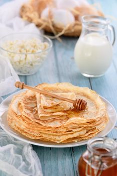 Homemade thin crepe (pancakes) with honey stacked in a stack, on a table with a pot of milk, a bowl of cottage cheese and eggs in a basket. Country style food. Traditional Slavonian, pagan holiday (Maslinitsa)