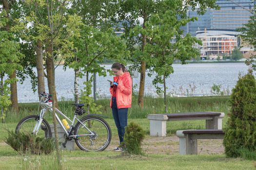 Belarus, Minsk - May 28, 2021: A woman with a bicycle is resting in the park and looks into a smartphone. Stock photography.