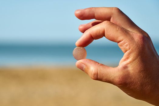 A pink piece of sea glass is held up with the sand, sea and sky in the background