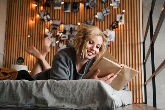 Happy Woman relaxing and reading book on cozy bed - Wooden wall and photos with lights - blurred background