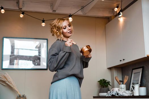 Smiling young woman in casual wear standing at the kitchen, holding ladle and whisk