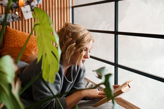 Blonde girl reading book on bed - cozy room with green plants