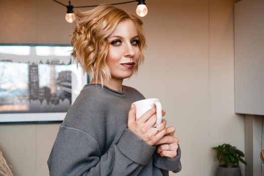 Close-up portrait of a beautiful woman with cup of coffee. Attractive and happy blonde in sweater drinks coffee. Stay at home - cozy house