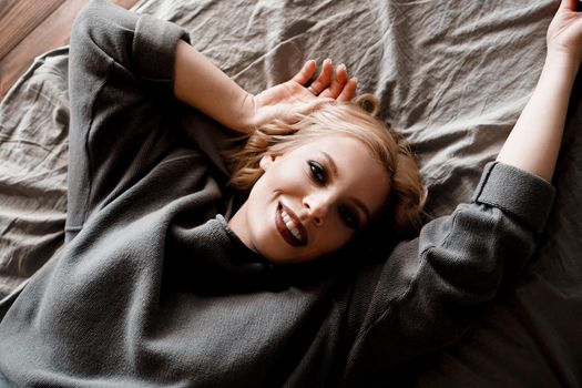 Beautiful young woman in gray sweater laughing lying in bed in her cozy bedroom. Cute girl relaxing at home. Happy morning