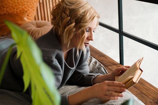 Blonde girl reading book on bed - cozy room with green plants