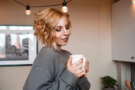 Close-up portrait of a beautiful woman with cup of coffee. Attractive and happy blonde in sweater drinks coffee. Stay at home - cozy house