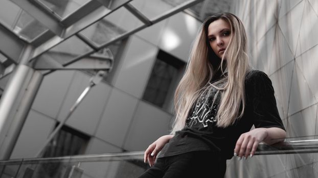 Urban style blonde girl with a stern look standing on the background of a modern building. Wearing black shirt. Girl power and subculture concept.